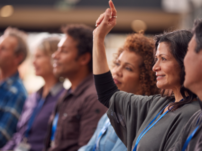 Adult learners engaged at a professional conference, one raising her hand to ask a question during a session