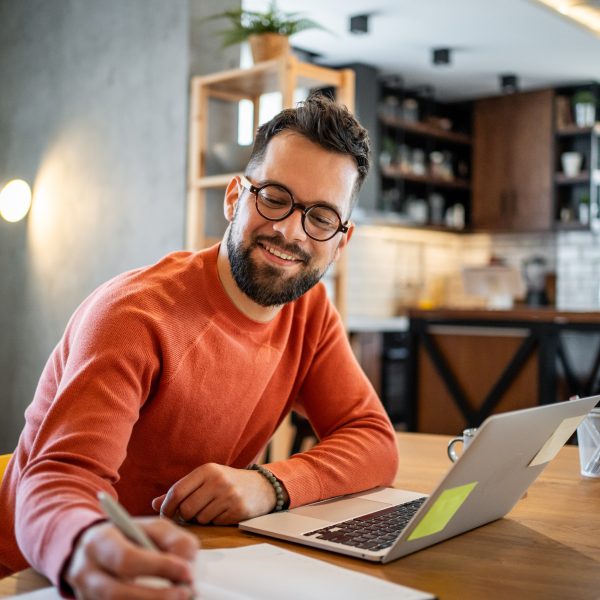 Young adult man working from home at his dining table, taking notes and smiling while engaging with his laptop. Enjoying a cozy atmosphere in his modern apartment, balancing work and study
