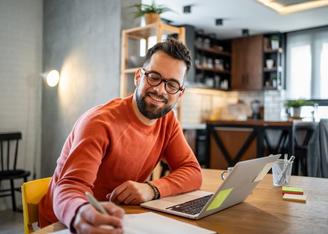 Young adult man working from home at his dining table, taking notes and smiling while engaging with his laptop. Enjoying a cozy atmosphere in his modern apartment, balancing work and study
